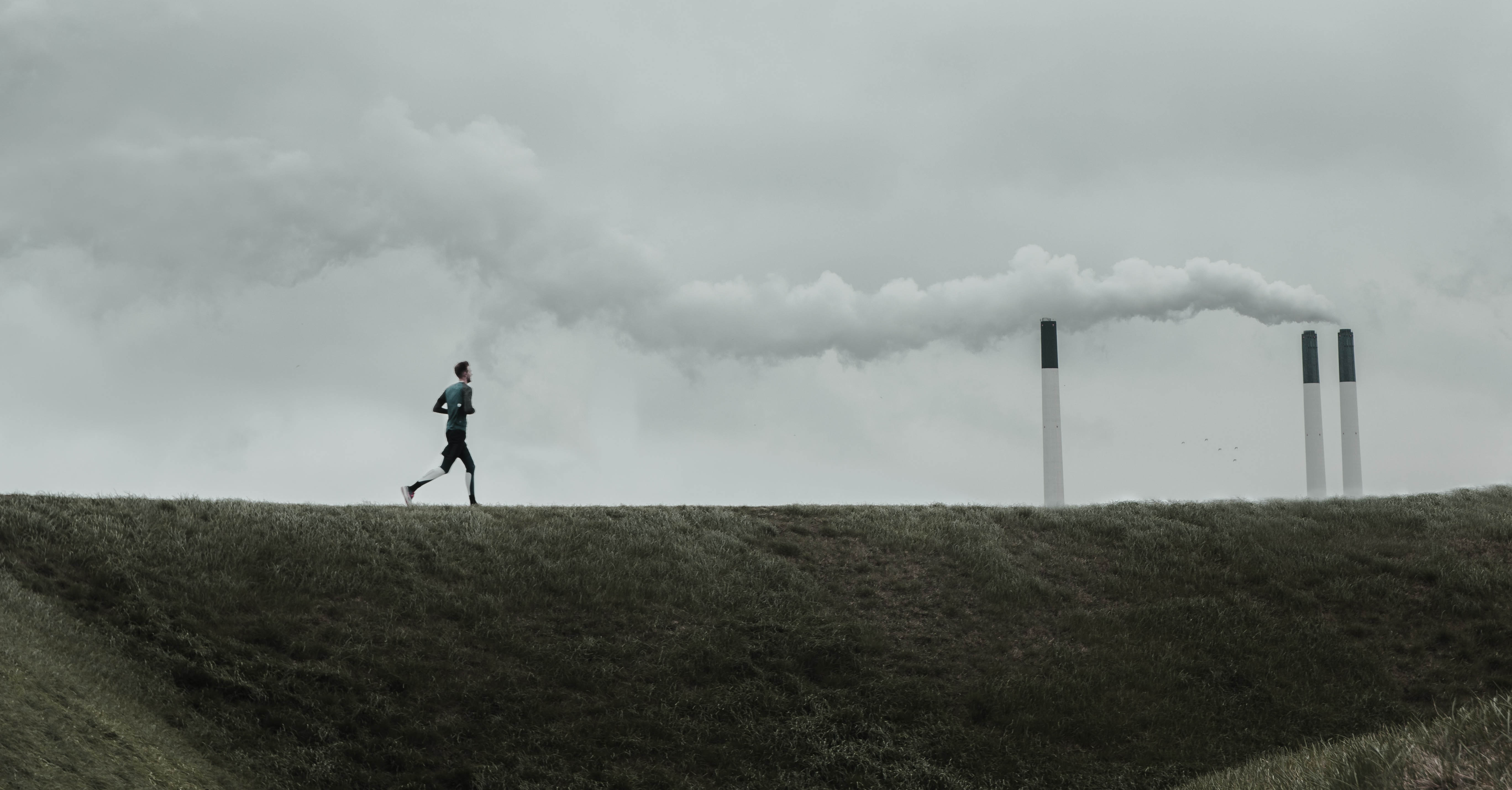 A person runs on a trail near a factory with a plume of smoke.