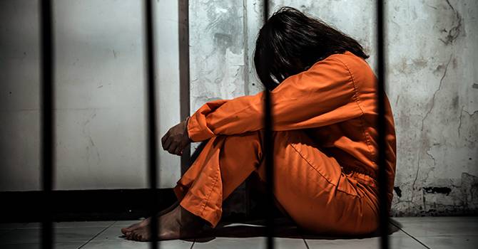 A woman in orange sits on the floor of a prison cell
